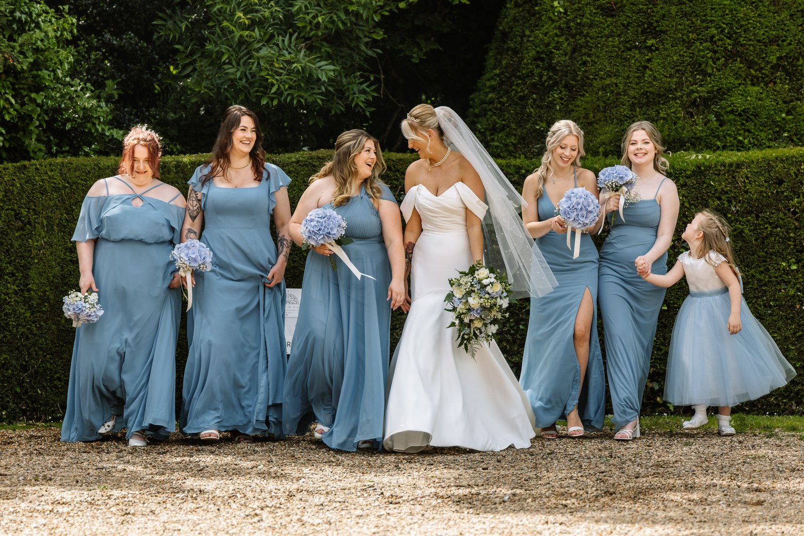 Bride and bridesmaids walking together past a tall clipped hedge