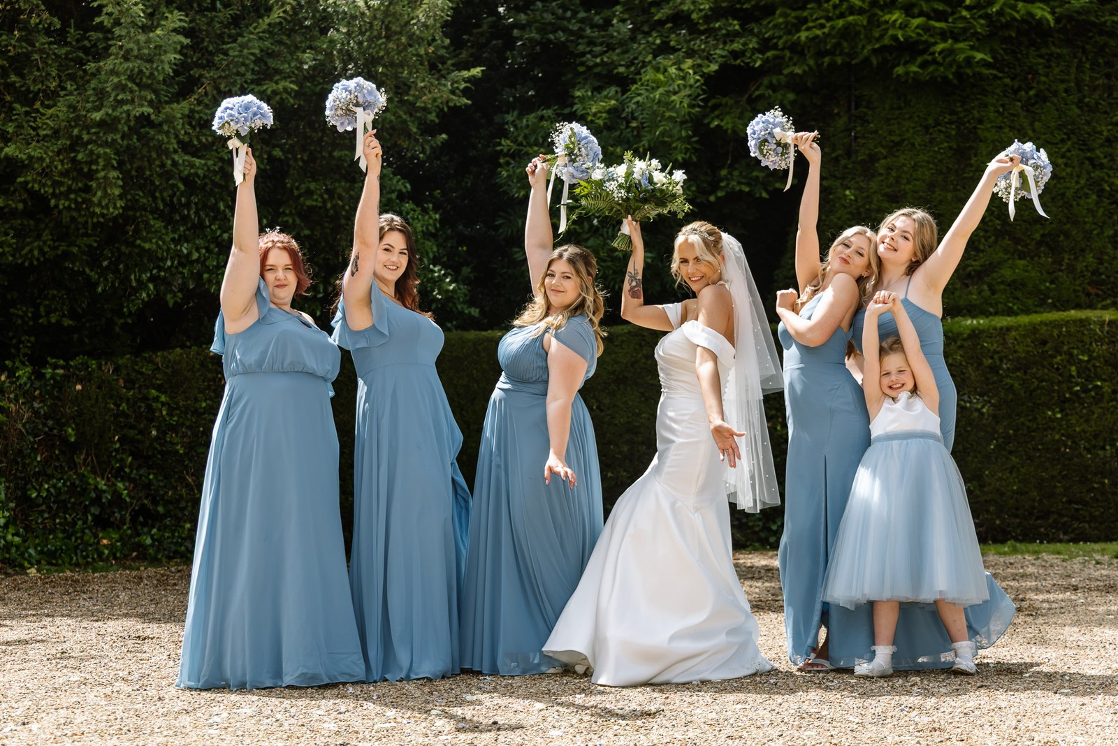 Bridesmaids cheering and raising their bouquets beside the clipped hedge