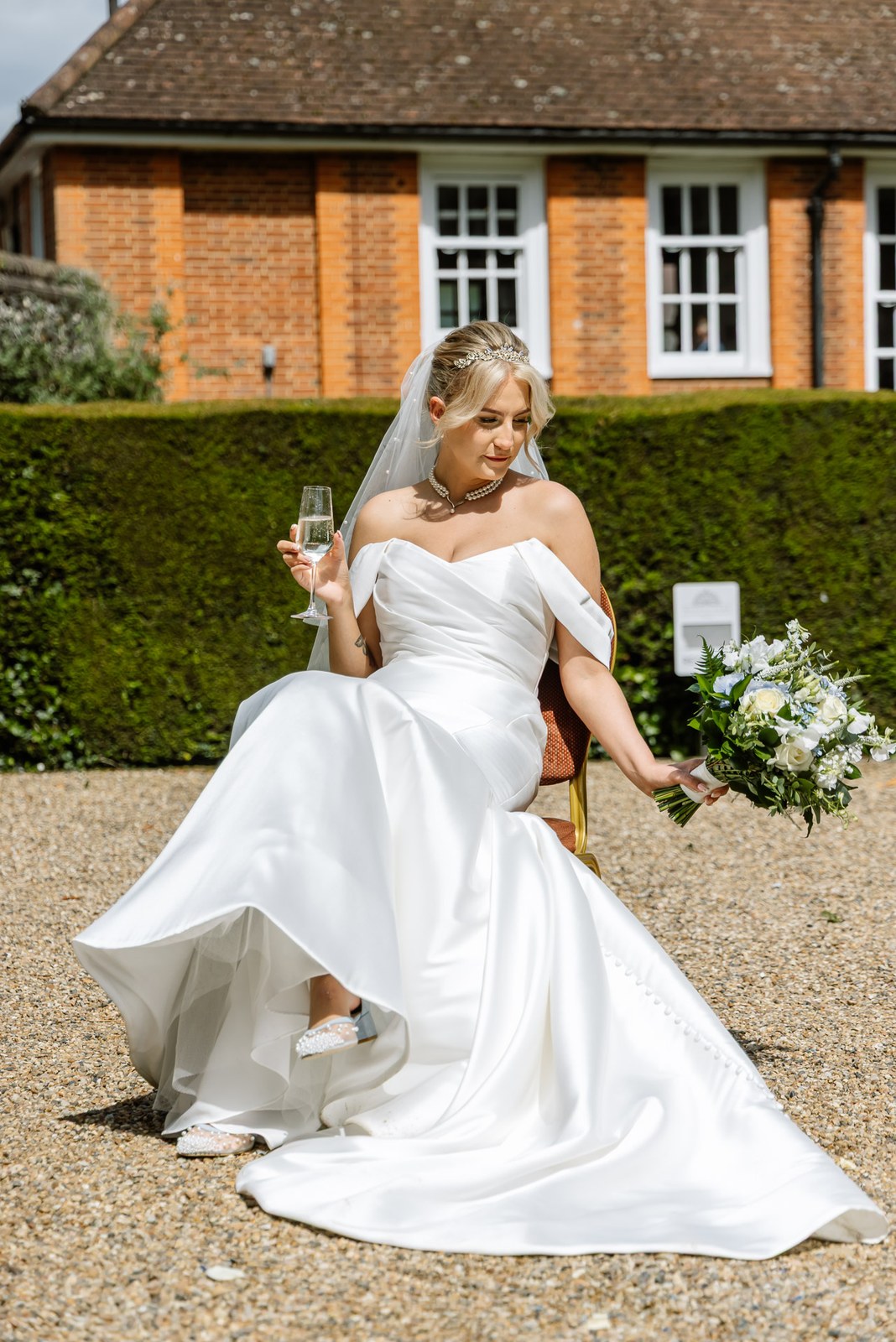The bride seated with a glass of champagne against a manicured hedge