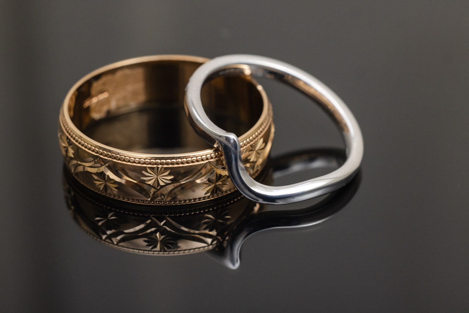 Close-up of the two wedding rings, a patterned gold band and a shaped white band, on a dark surface