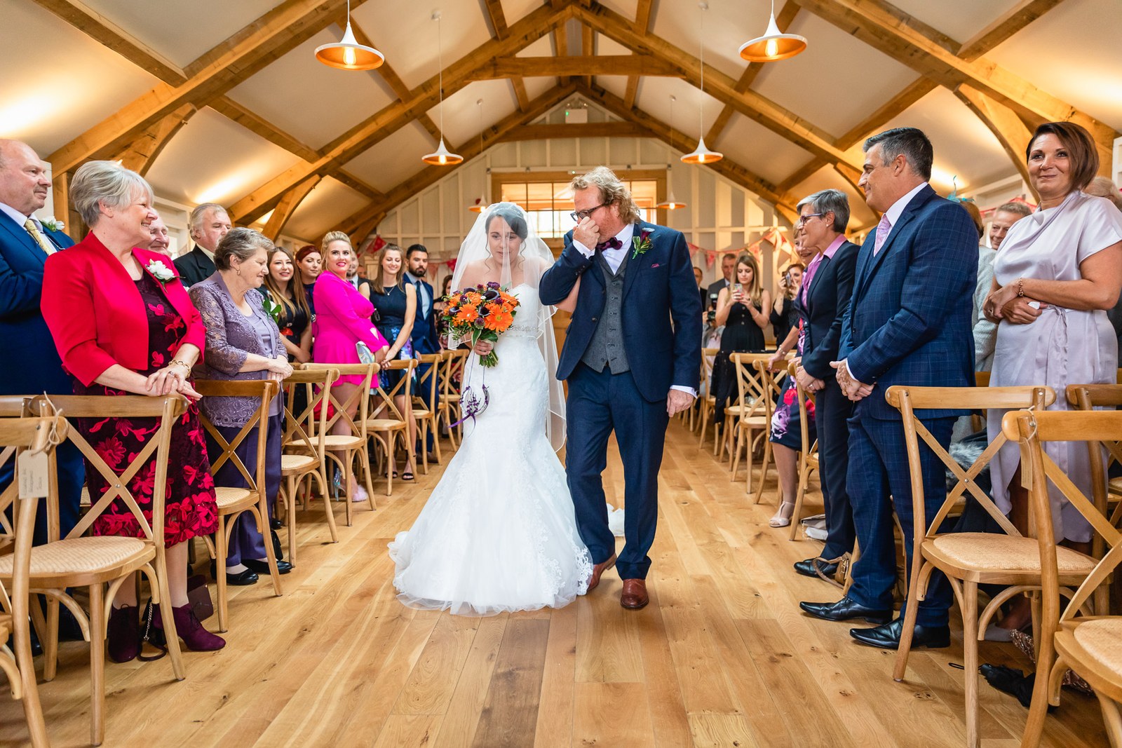 The bride walking down the aisle on her escort's arm through the timbered barn