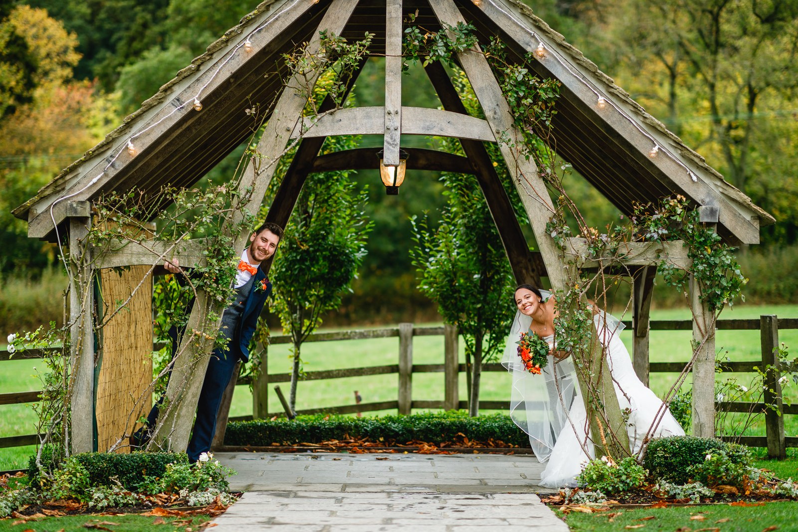 The groom leaning on the oak arbour, smiling across at the bride
