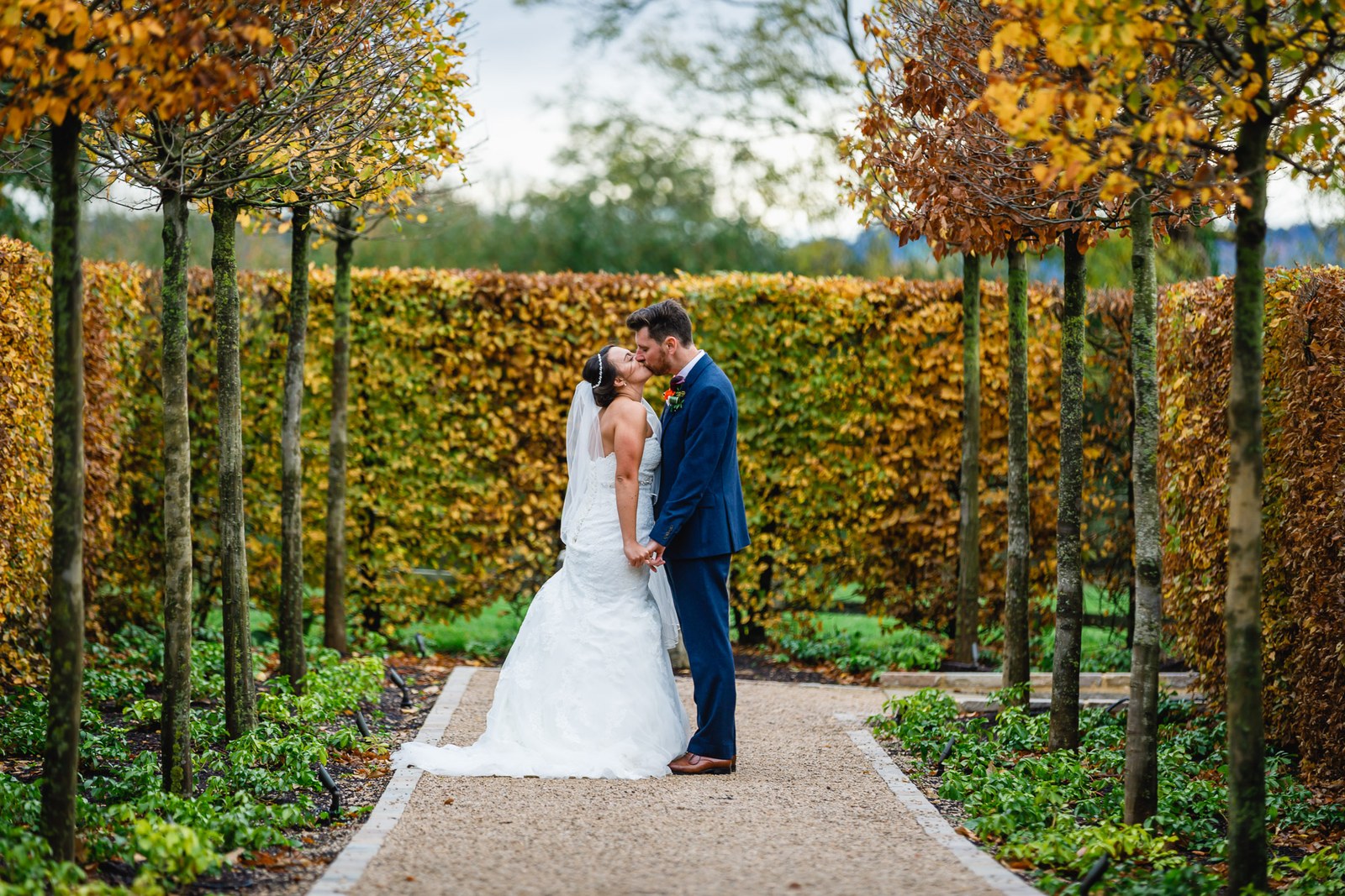 The newlyweds kissing in the avenue of pleached trees in autumn colour at Hyde House