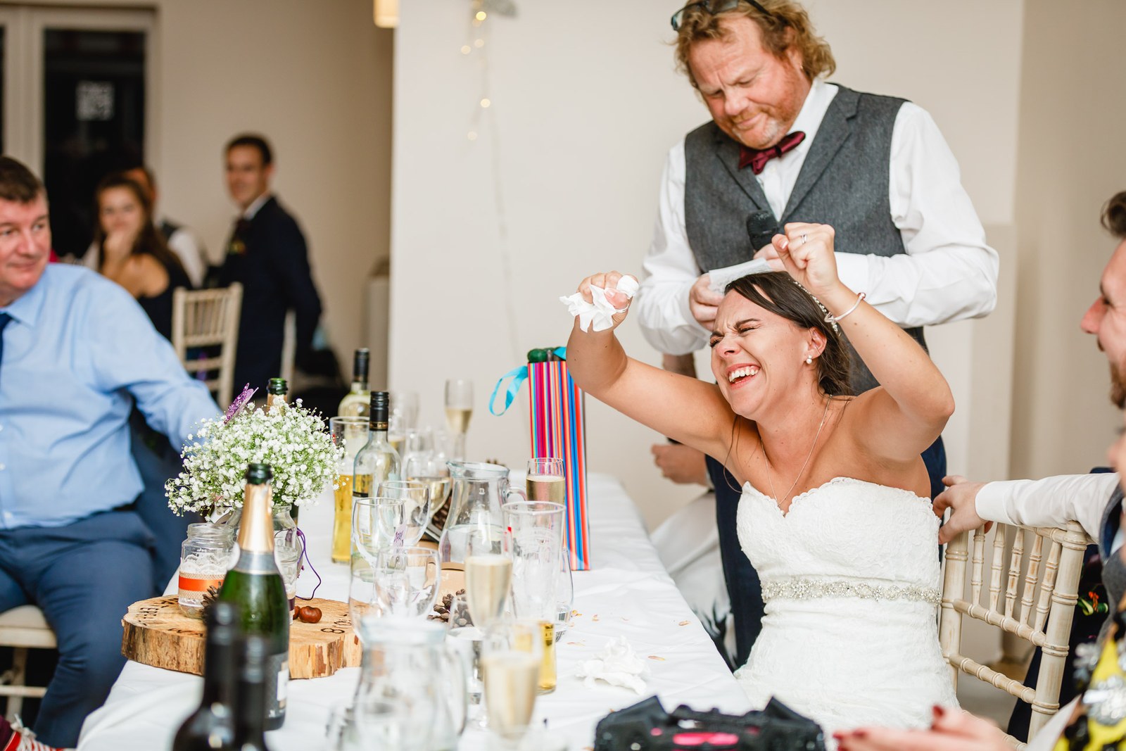 The bride laughing as a guest holds a napkin over her head at the top table