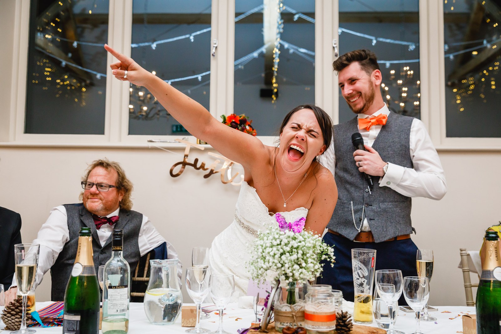 The bride cheering with a raised arm as the groom speaks into the mic