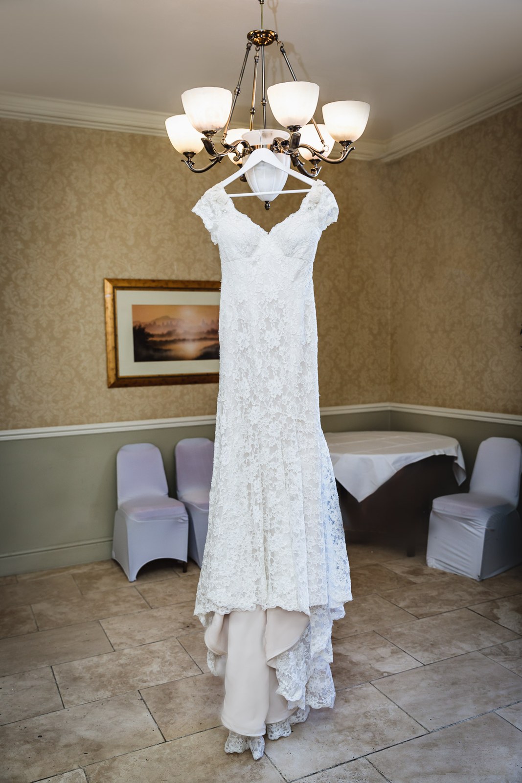 Bride's lace wedding dress on a hanger below a chandelier in a room with covered chairs