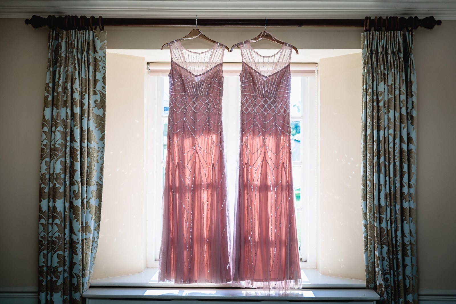 Two beaded rose-pink bridesmaid dresses hanging in a bright window with patterned curtains