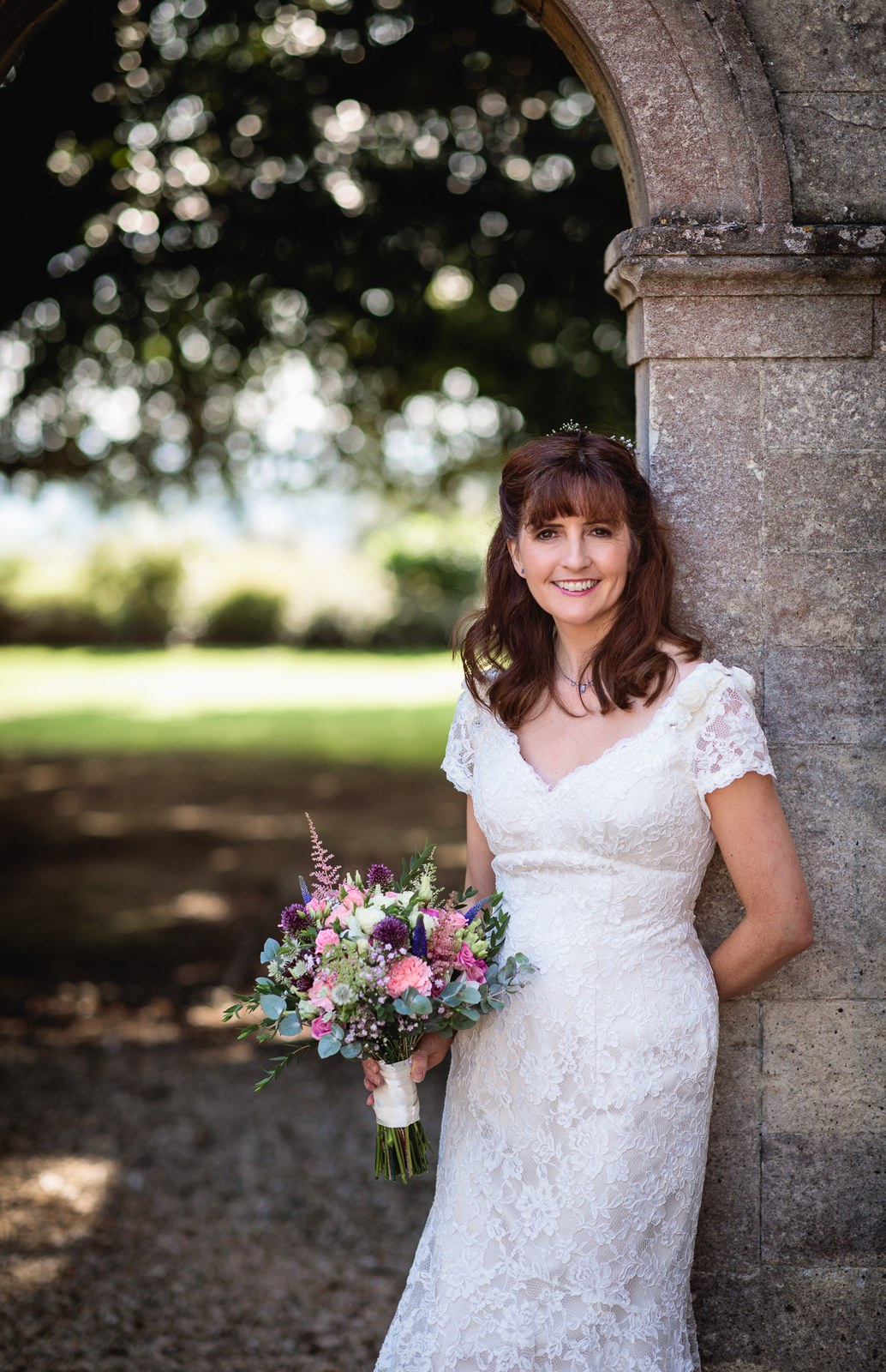 Bride holding her bouquet, leaning against a stone archway beneath a tree