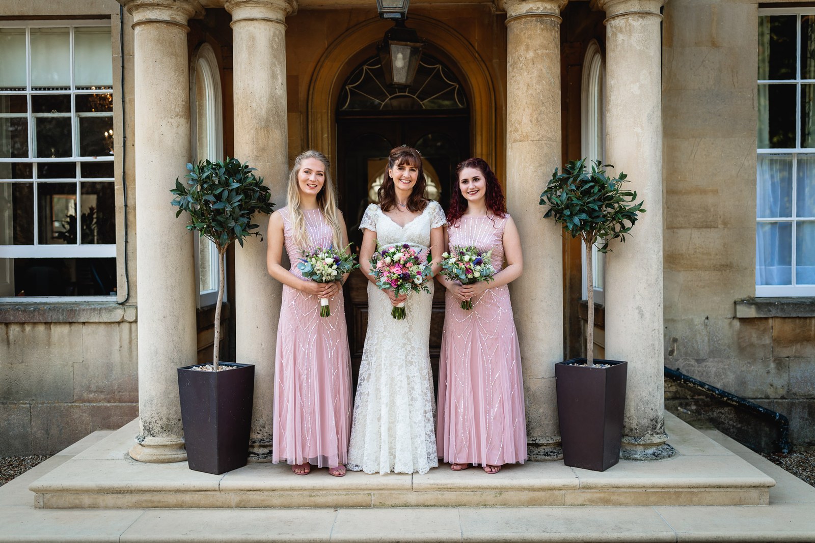 Bride and two pink bridesmaids between the stone columns of the hotel entrance