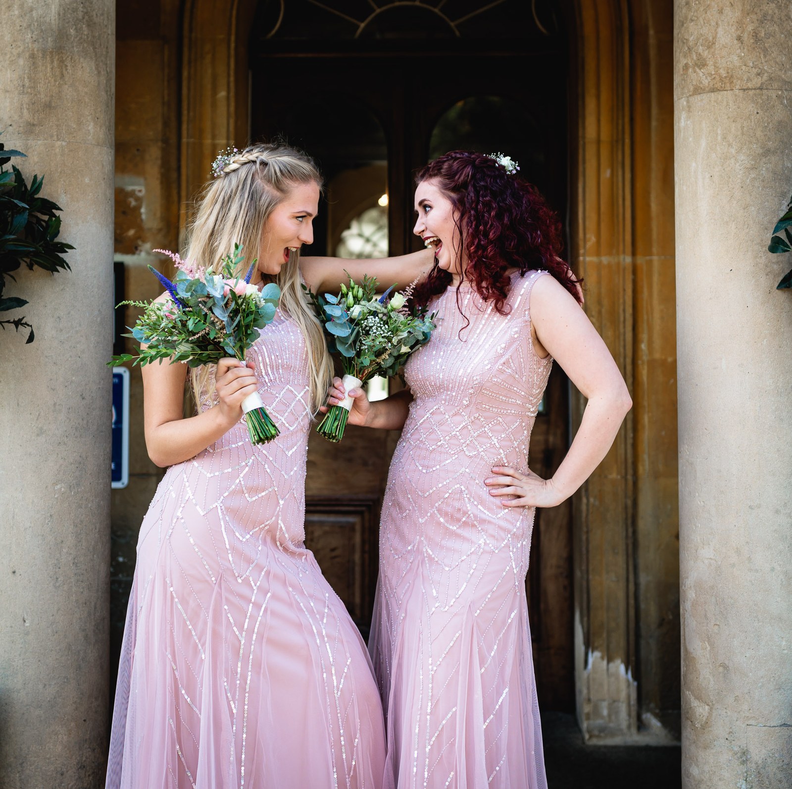 Two bridesmaids in beaded pink gowns laughing together under a stone portico