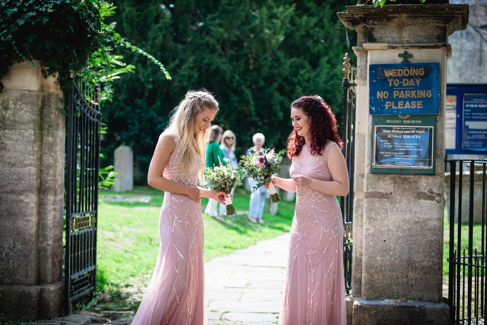 Two bridesmaids adjusting flowers at a churchyard gate by a wedding-today sign