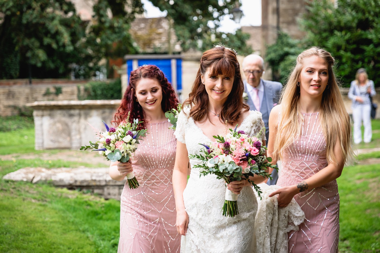 Bride walking across the churchyard with two bridesmaids, a guest following behind