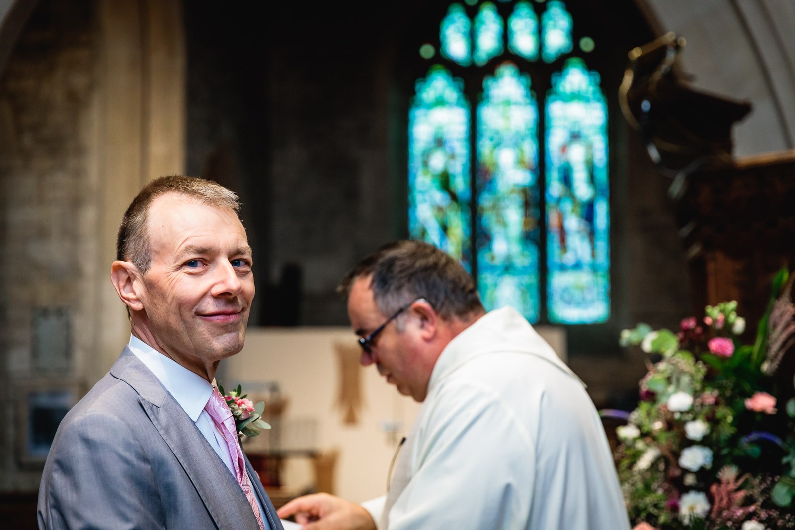 Groom smiling beside the vicar at the front of the church, stained glass behind