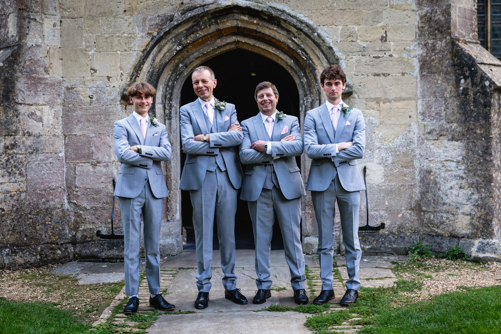 The groom and three groomsmen in grey suits, arms folded, beneath a pointed stone church archway