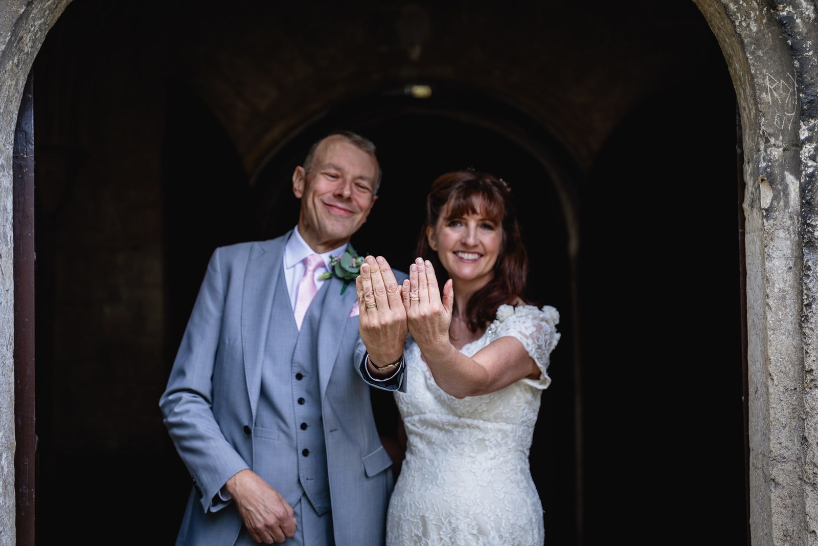 Bride and groom holding up their wedding rings to the camera in a stone doorway