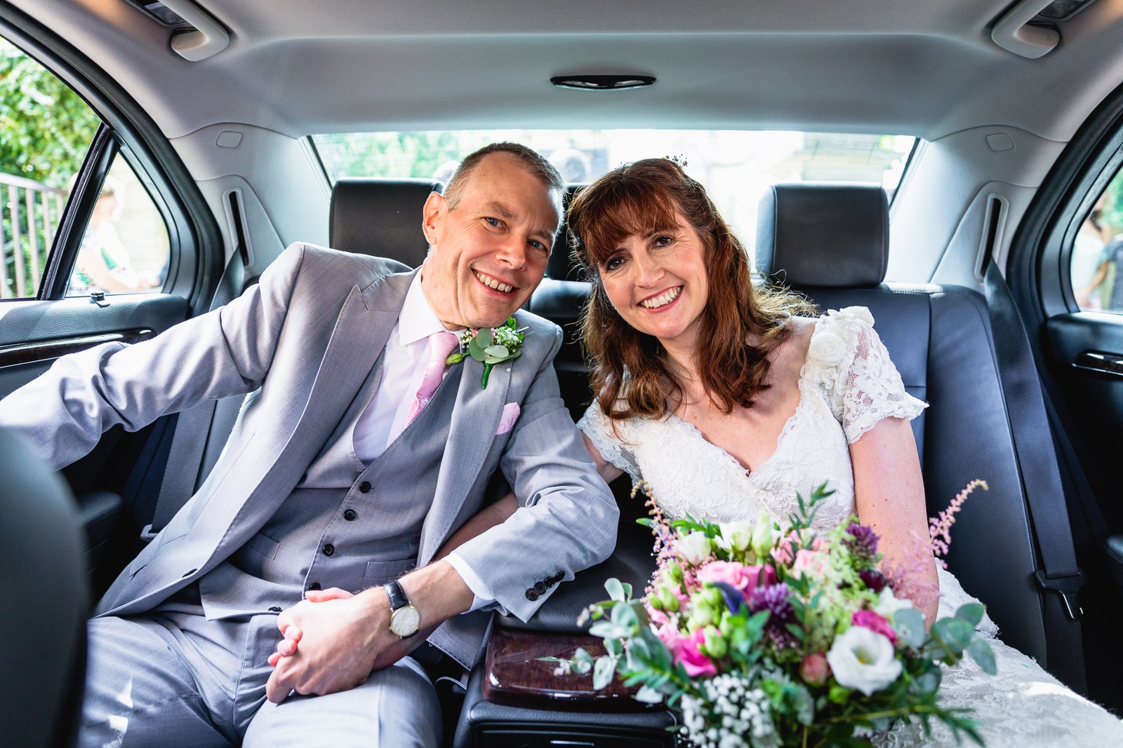 Bride and groom seated together in the back of the wedding car, bouquet on her lap