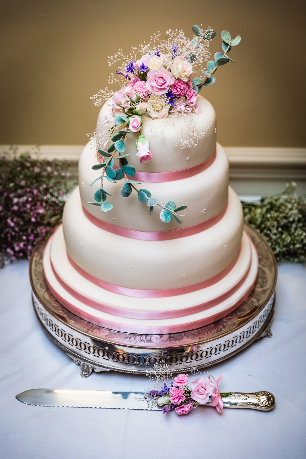 Three-tier wedding cake trimmed with pink ribbon and fresh flowers, a knife alongside