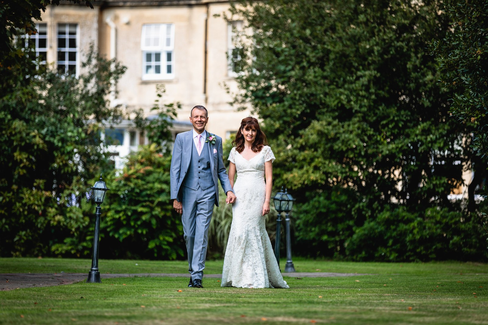 The couple crossing the lawn with the honey-stone hotel behind them