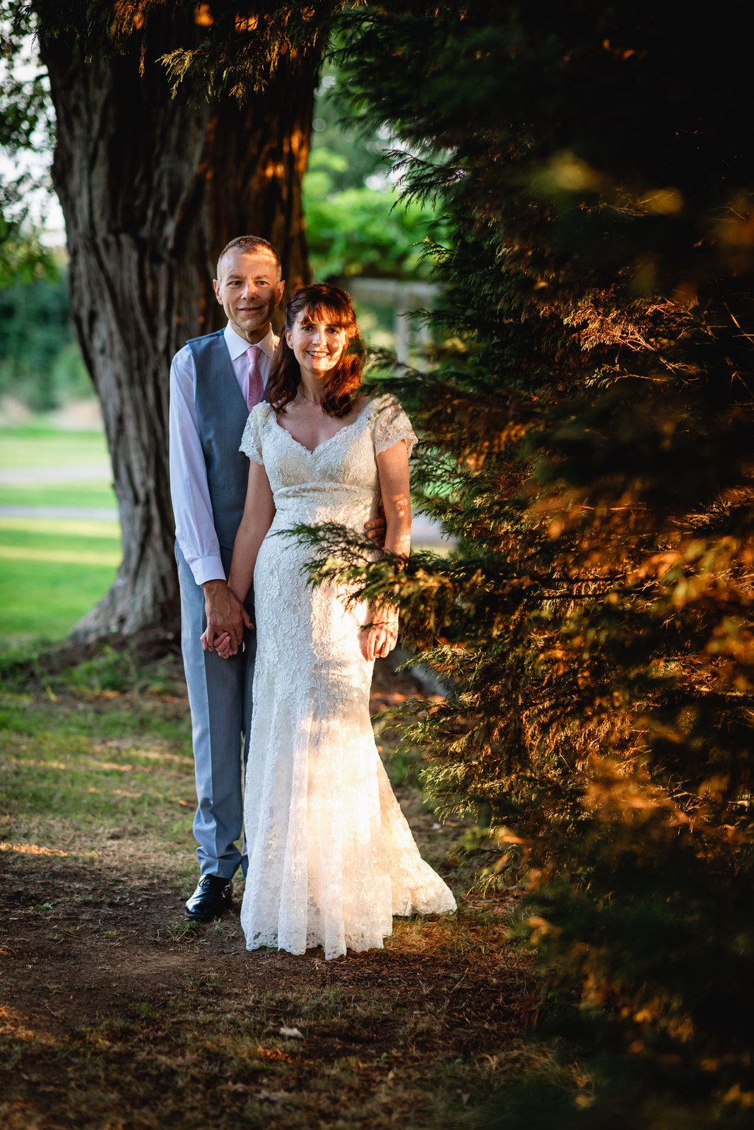 The couple standing beneath a large tree in the grounds at golden hour
