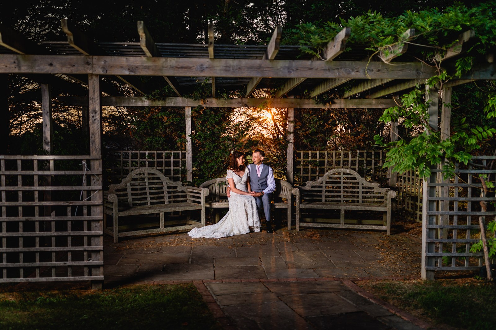 The couple on a wooden bench beneath a wisteria-clad pergola at sunset