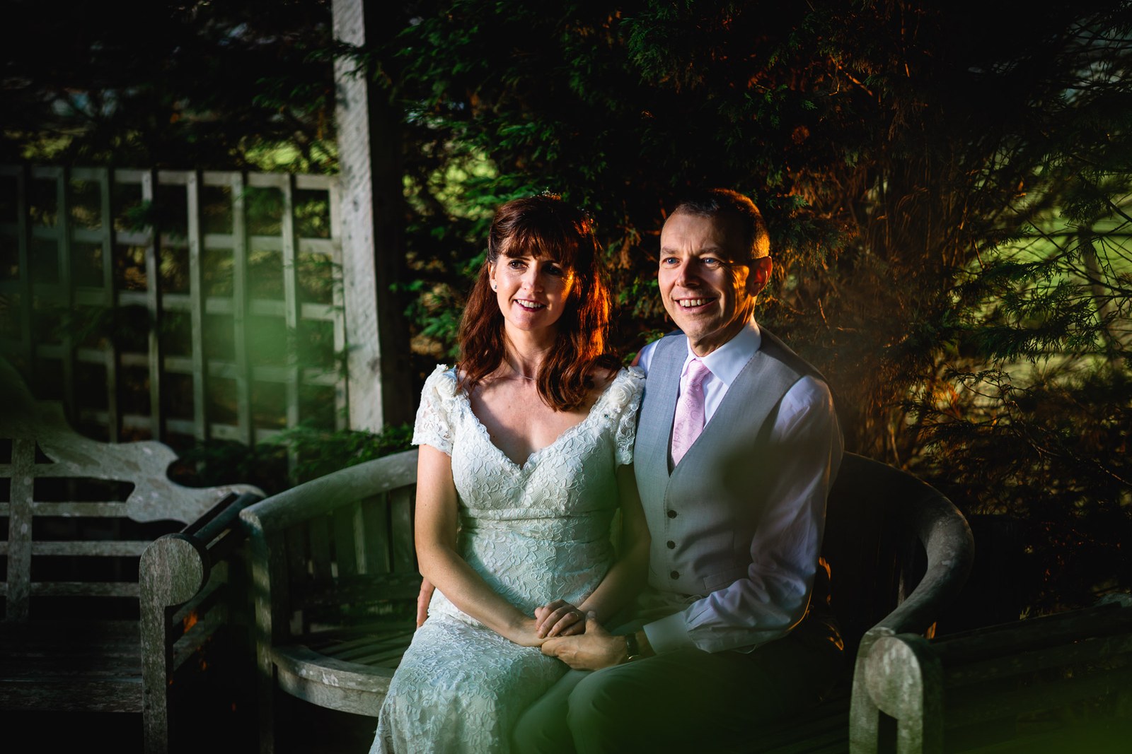 Bride and groom by a garden bench in the grounds in warm evening light