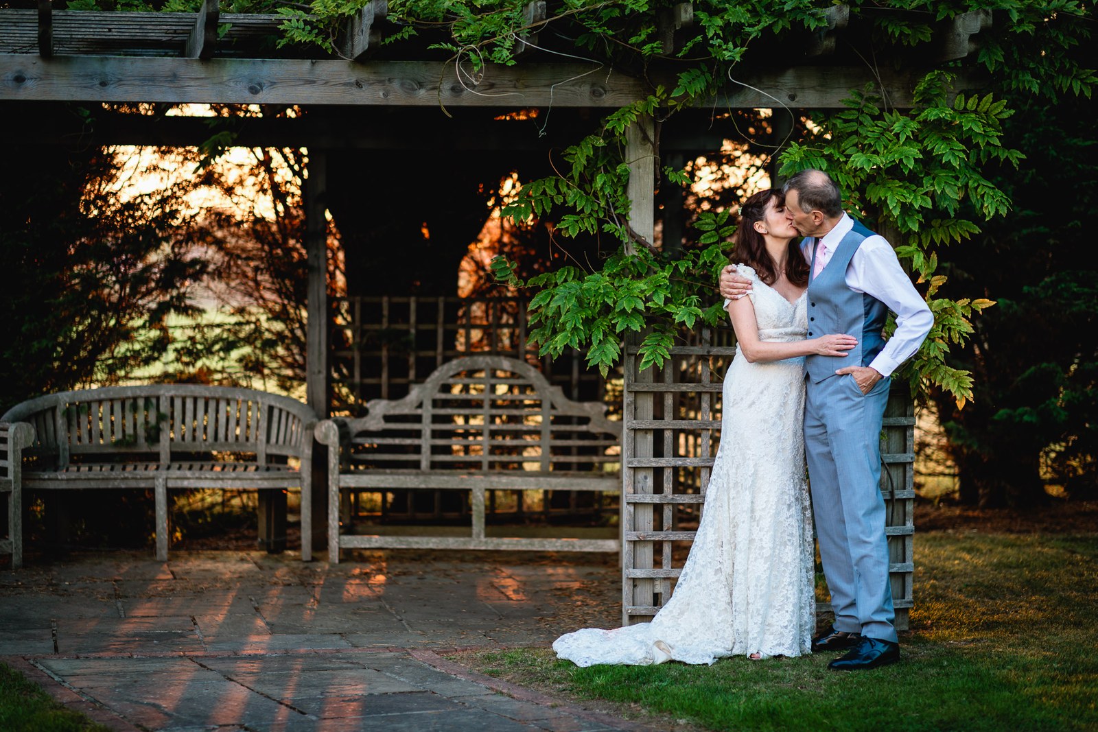 The couple kissing under the pergola at sunset beside garden benches