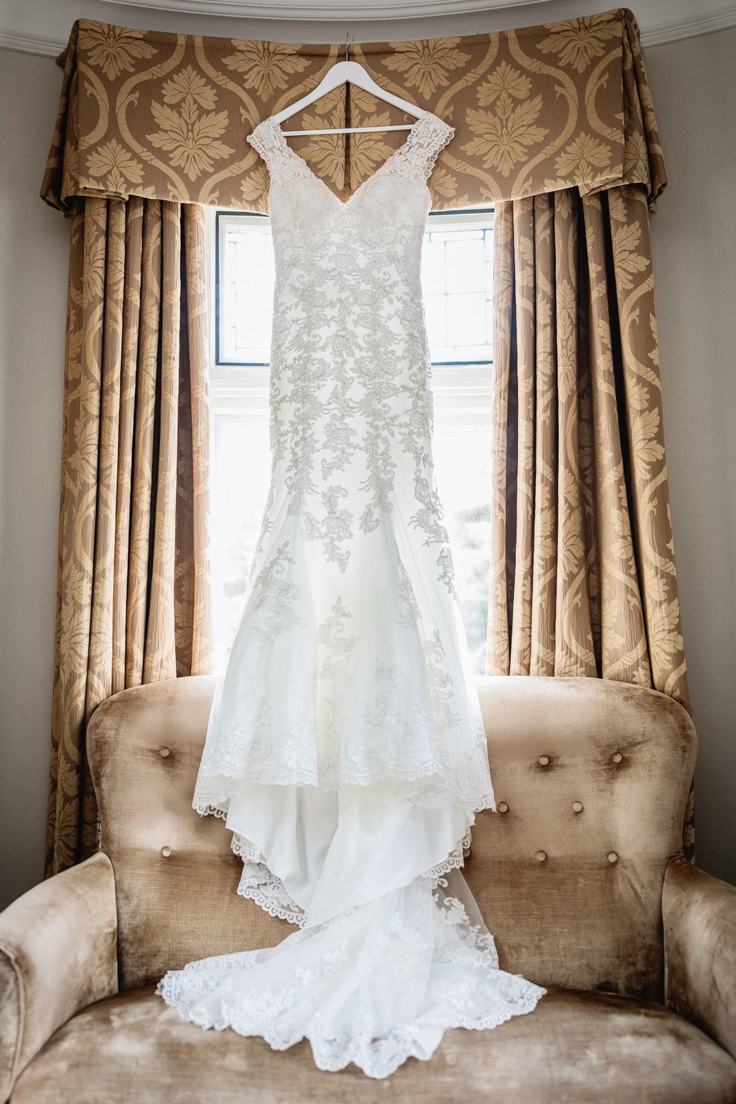 Lace wedding dress on a hanger in the window of a Macdonald Berystede Hotel suite, gold damask curtains