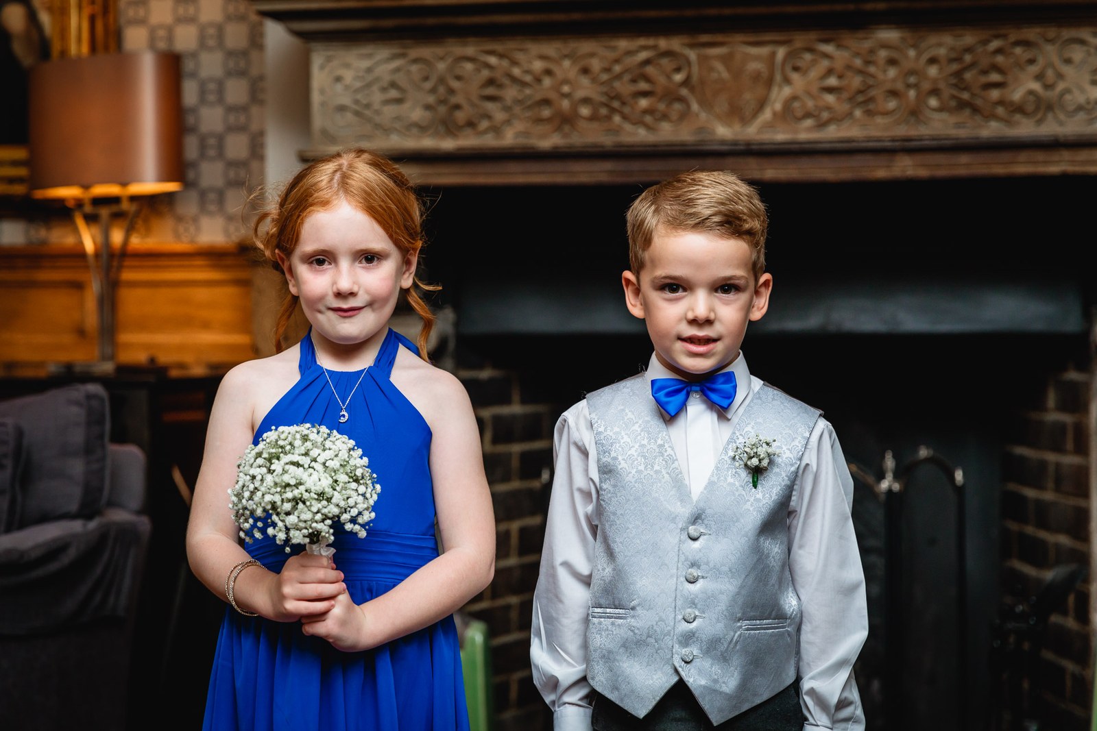 A flower girl with a posy and a page boy in grey waistcoat and blue bow tie by a fireplace
