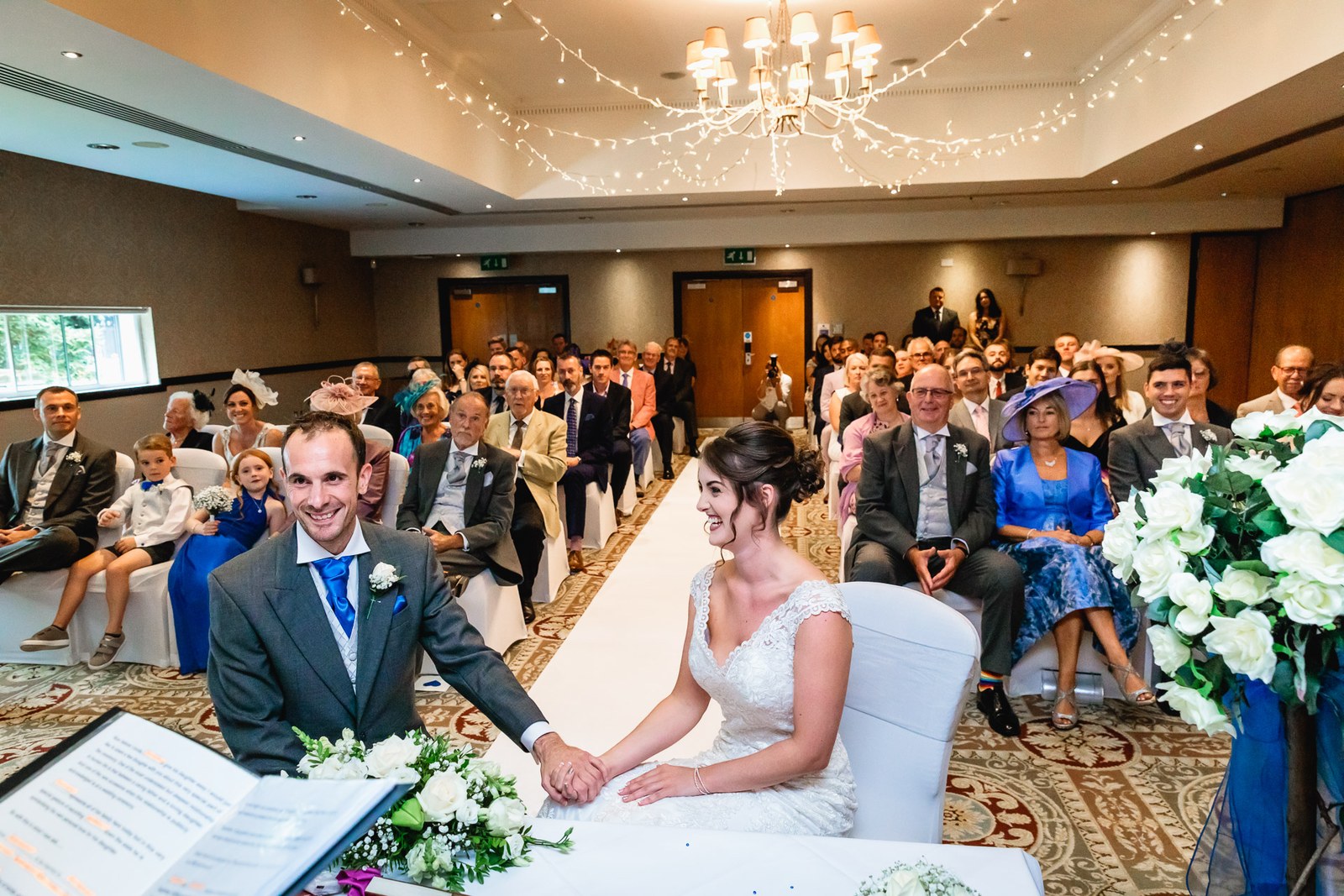 Bride and groom seated during the ceremony beneath a chandelier and fairy lights