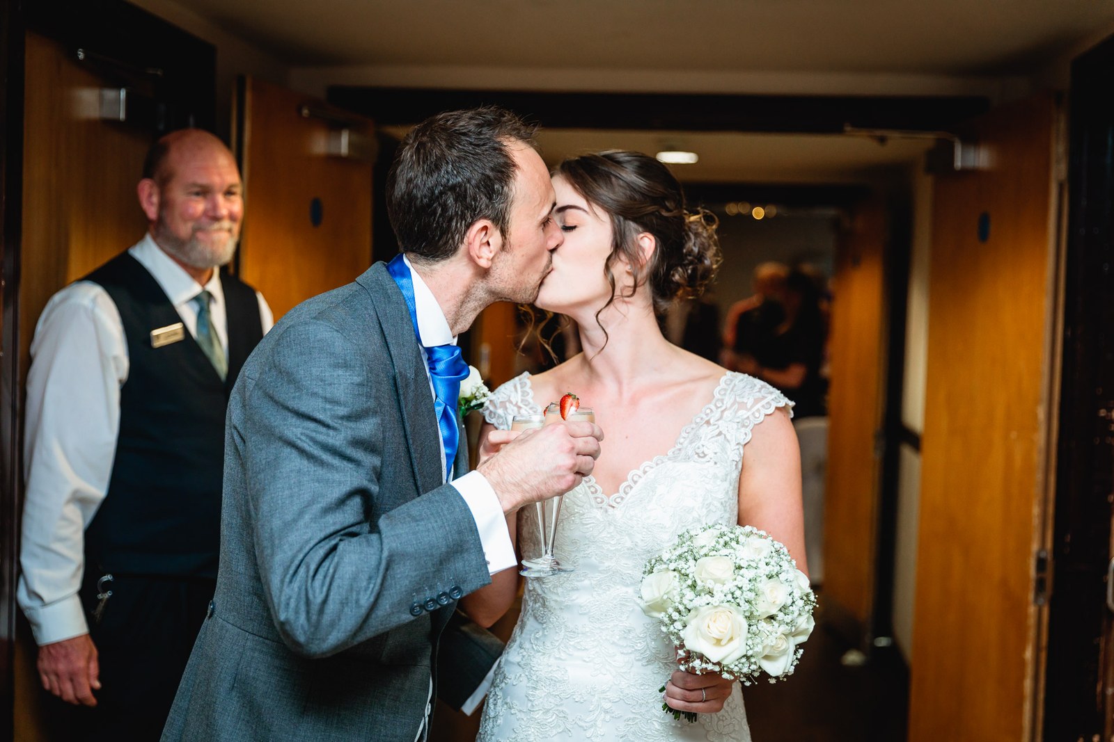 Bride and groom kissing while holding their drinks, a staff member behind