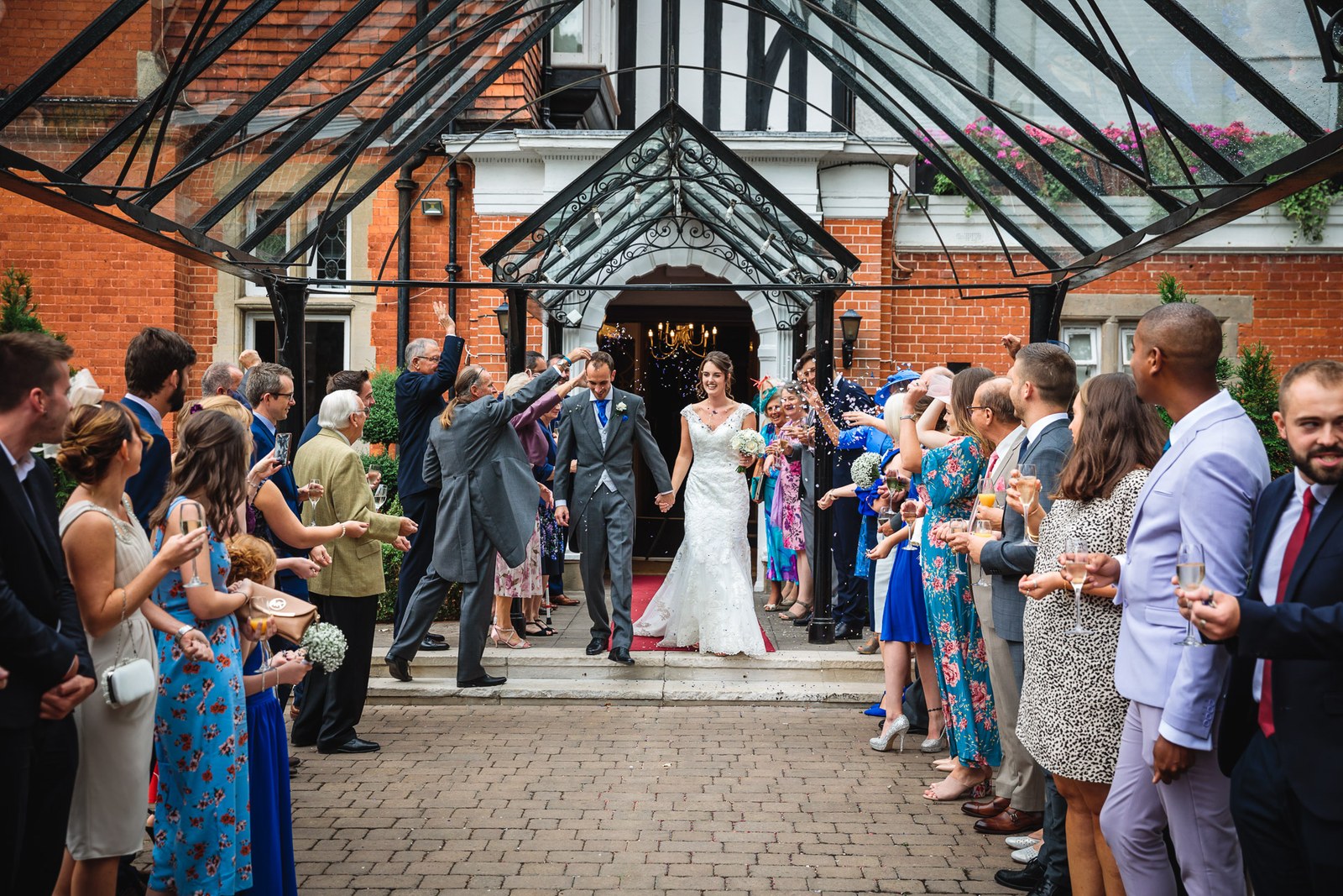 The couple leaving through a confetti line under the glass and timber porch of Macdonald Berystede Hotel