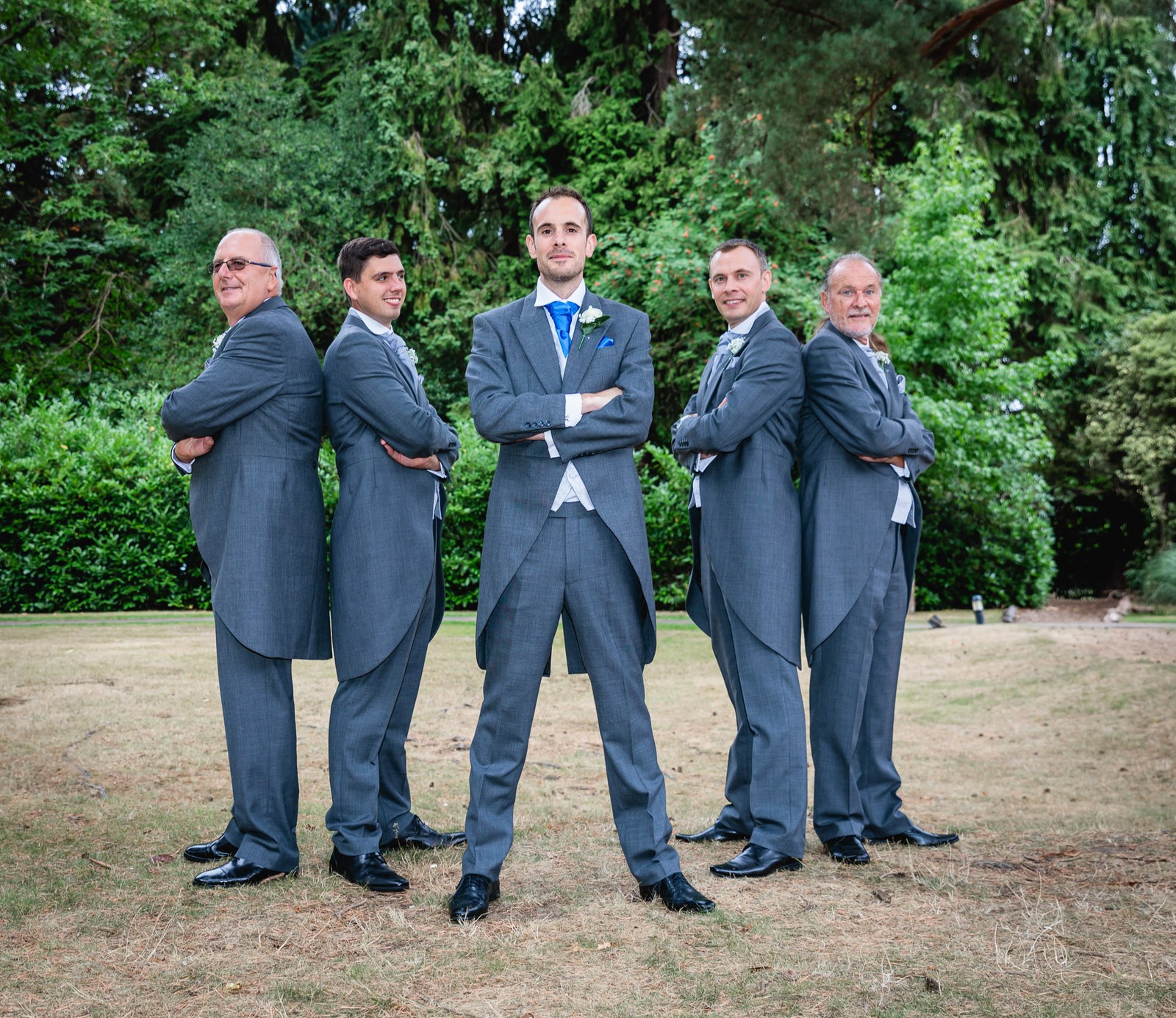 Groom and groomsmen standing back to back in a line, arms folded, on the lawn
