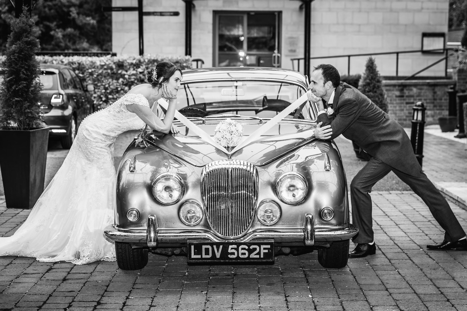 Black and white of the couple leaning on the bonnet of their classic wedding car
