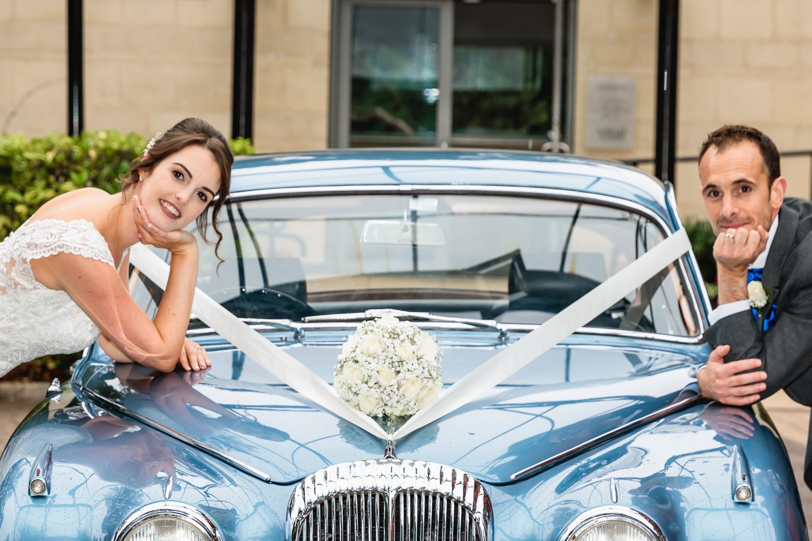 Bride and groom leaning on the ribbon-decorated blue classic car