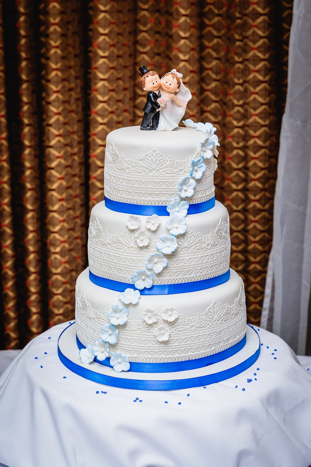 Three-tier white wedding cake with blue ribbon, flowers and a bride-and-groom topper