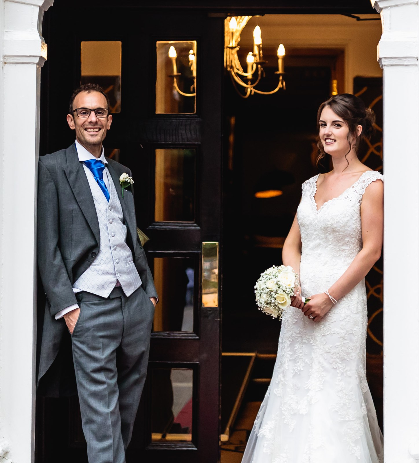 Bride and groom either side of the open panelled front door, chandelier glowing inside
