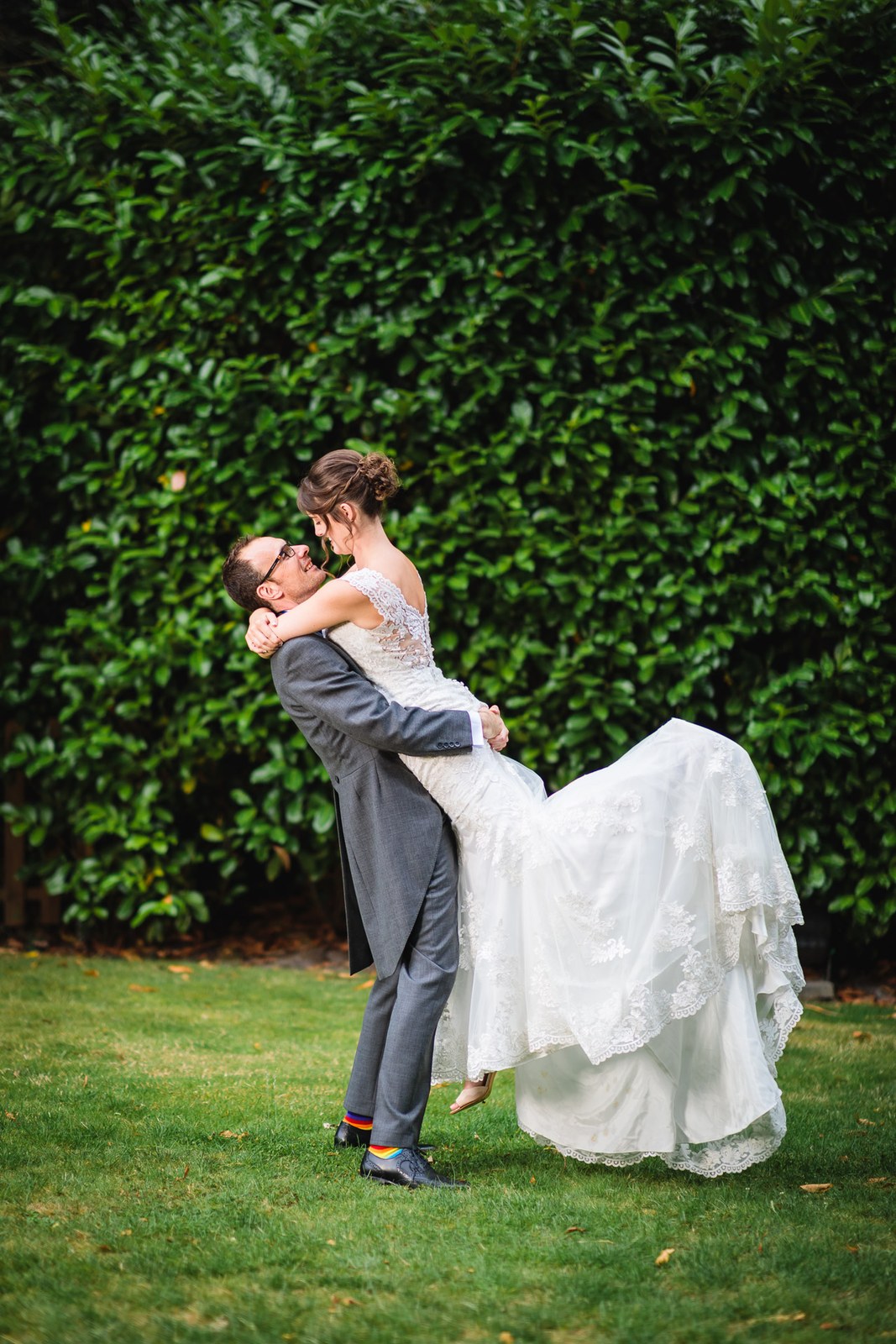 Groom lifting the bride in his arms against a tall green hedge in the grounds
