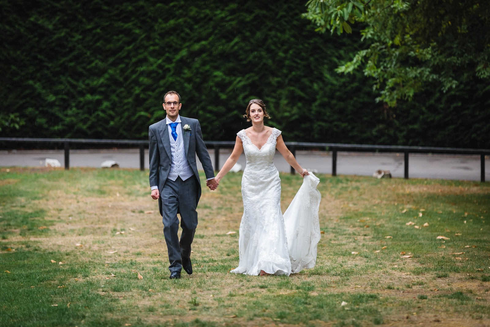 Bride and groom walking hand in hand across the lawn, hedge behind