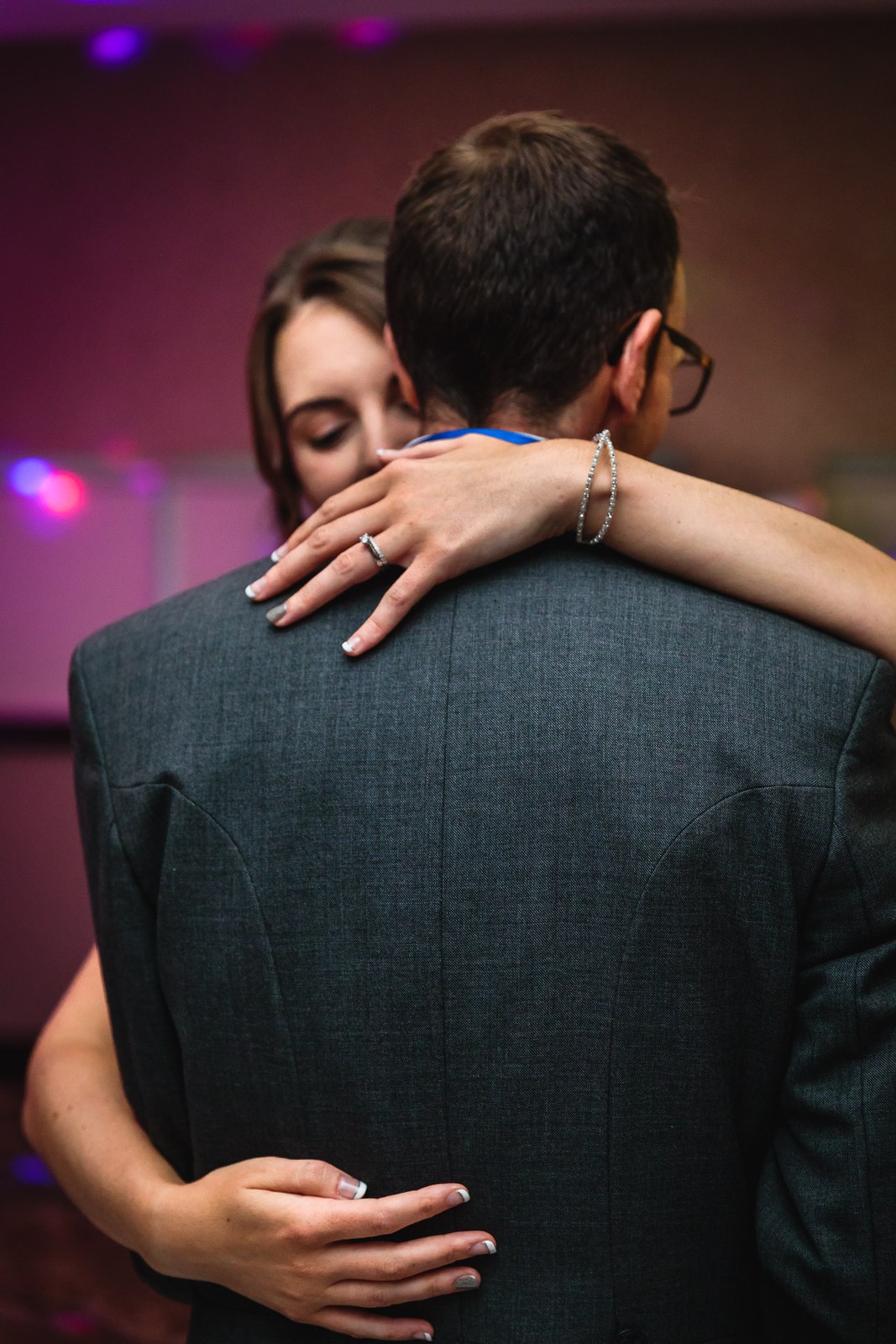 Bride embracing the groom, seen from behind, her ringed hand on his shoulder, purple light