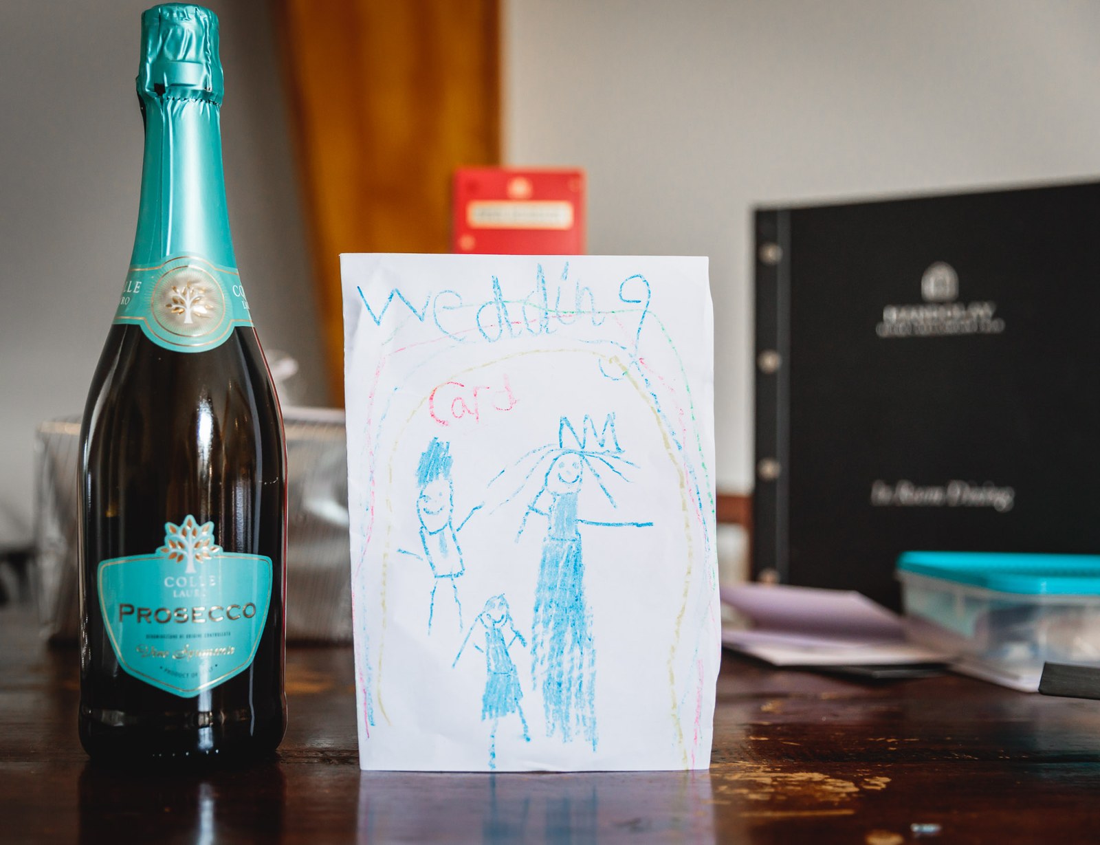 A bottle of Prosecco beside a child's crayon drawing of the wedding