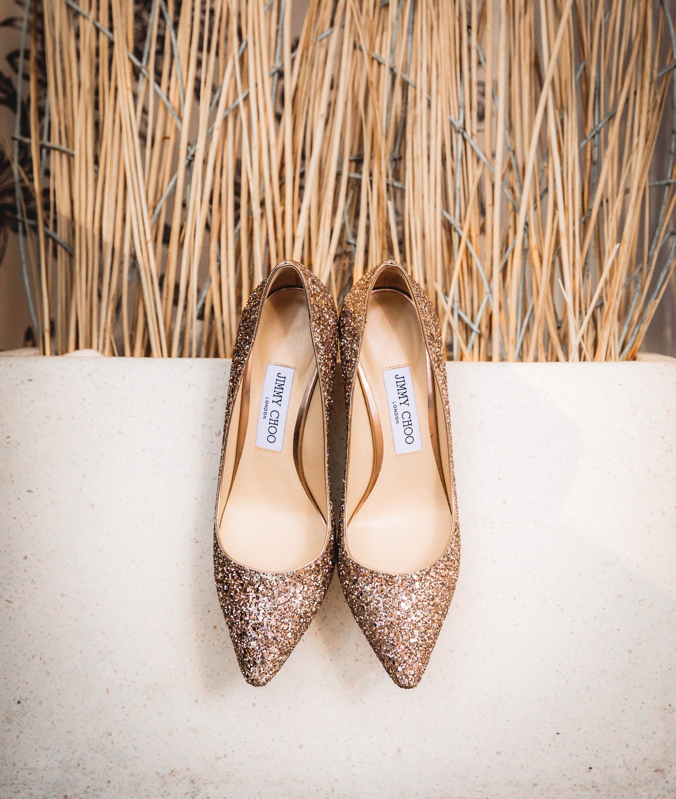 A pair of glittering rose-gold heels resting against a screen of reeds