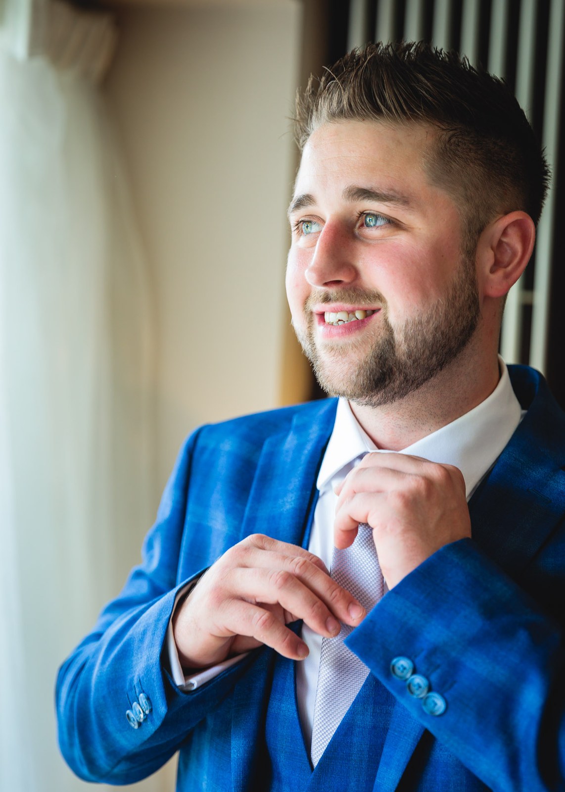 Groom in a bright blue suit adjusting his tie by a window