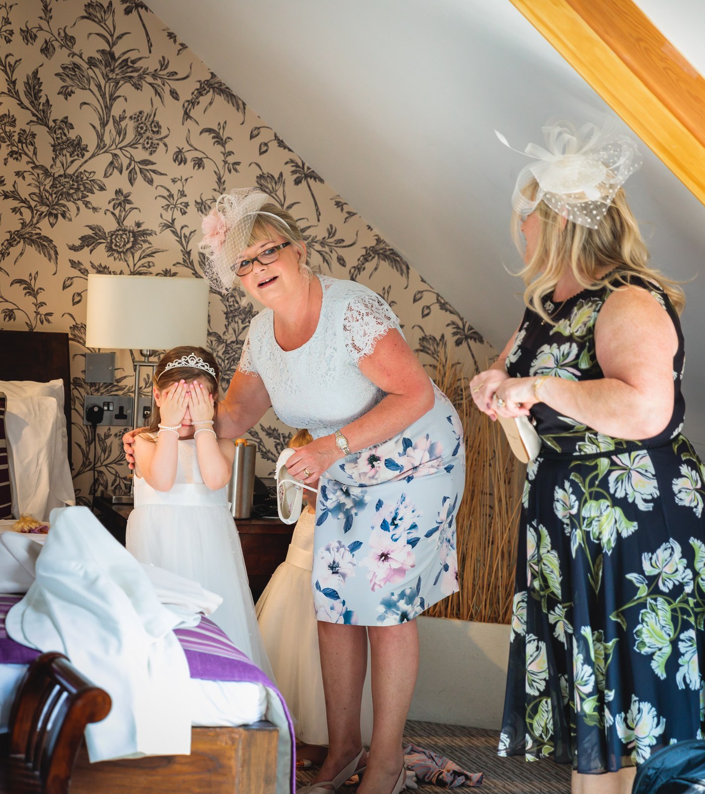 An older guest helping a giggling flower girl get ready in a floral room