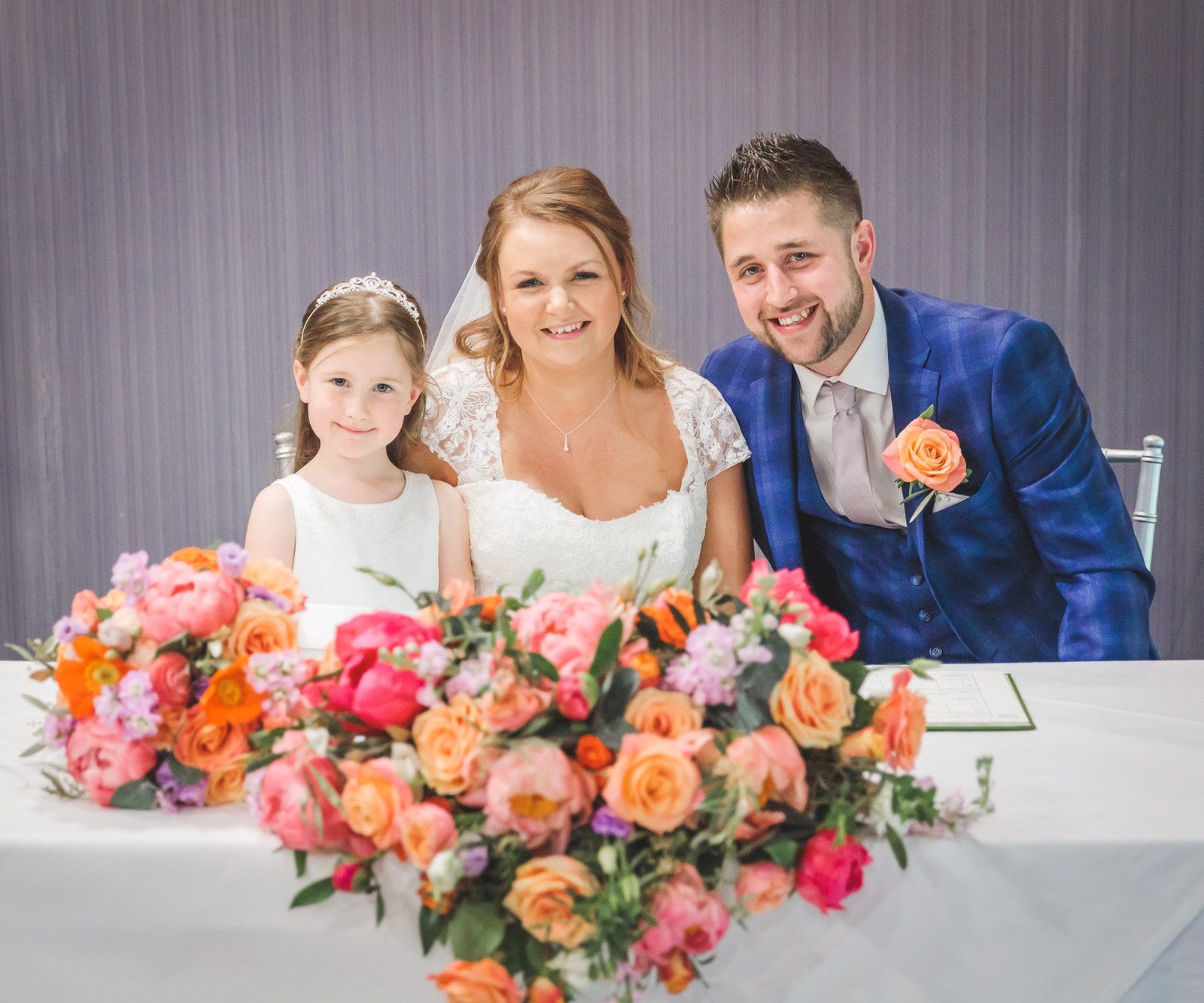 Bride, groom and flower girl posing behind the signing table of bright flowers