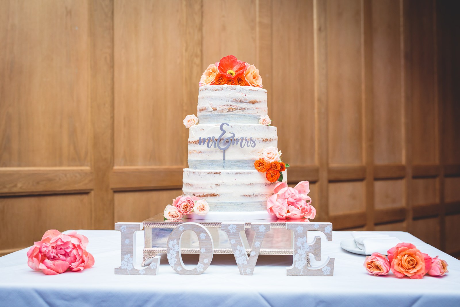 A semi-naked tiered wedding cake with fresh flowers beside wooden LOVE letters