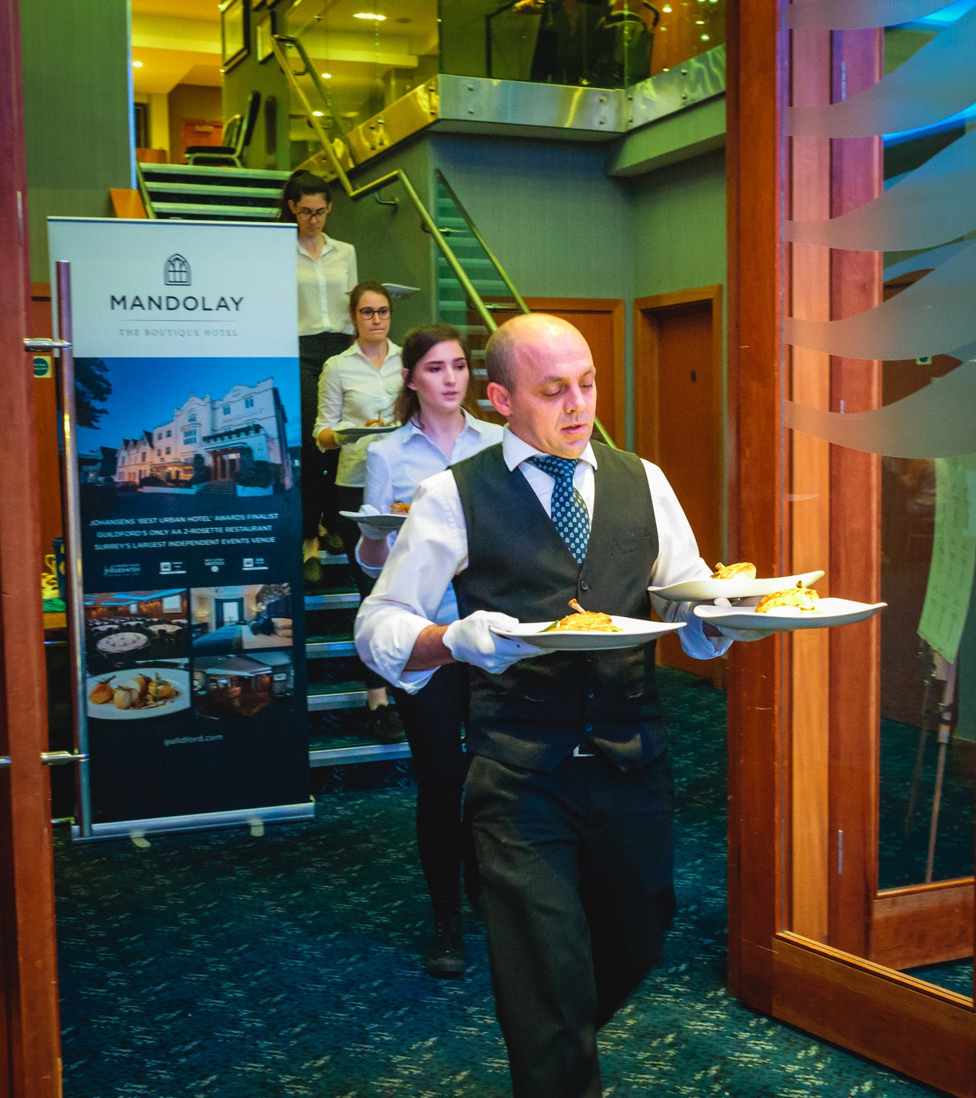A waiter carrying plated meals past a Mandolay Hotel banner