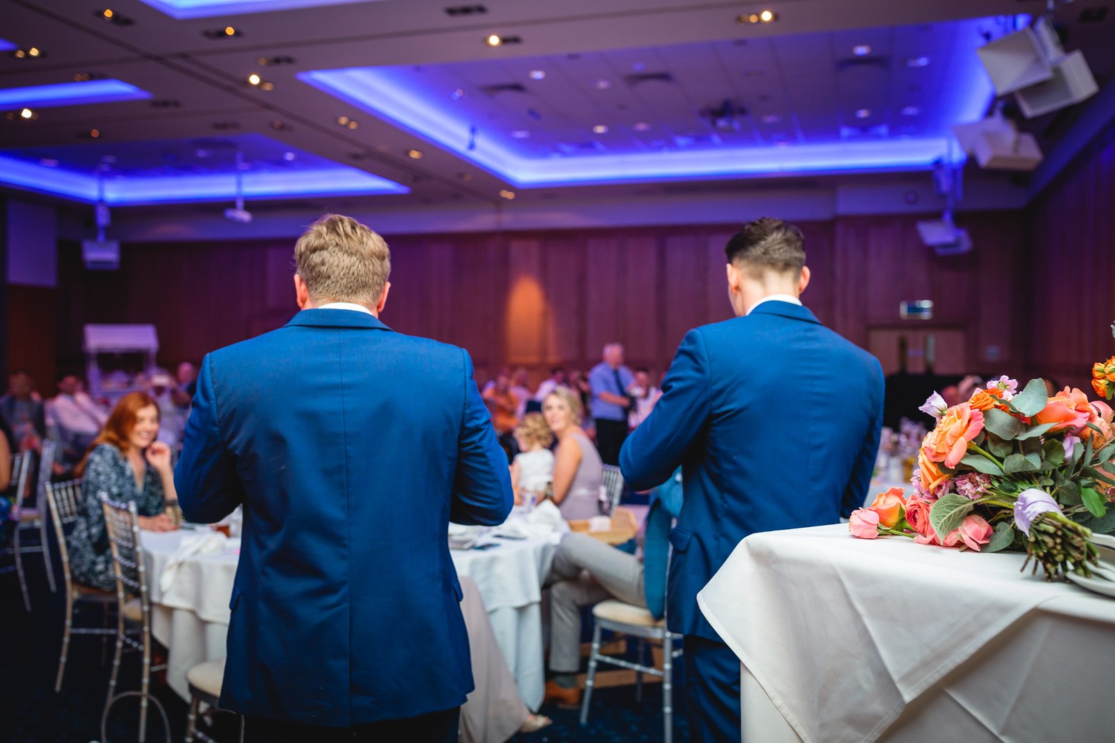 Two groomsmen mingling among the reception tables, a bouquet in the foreground