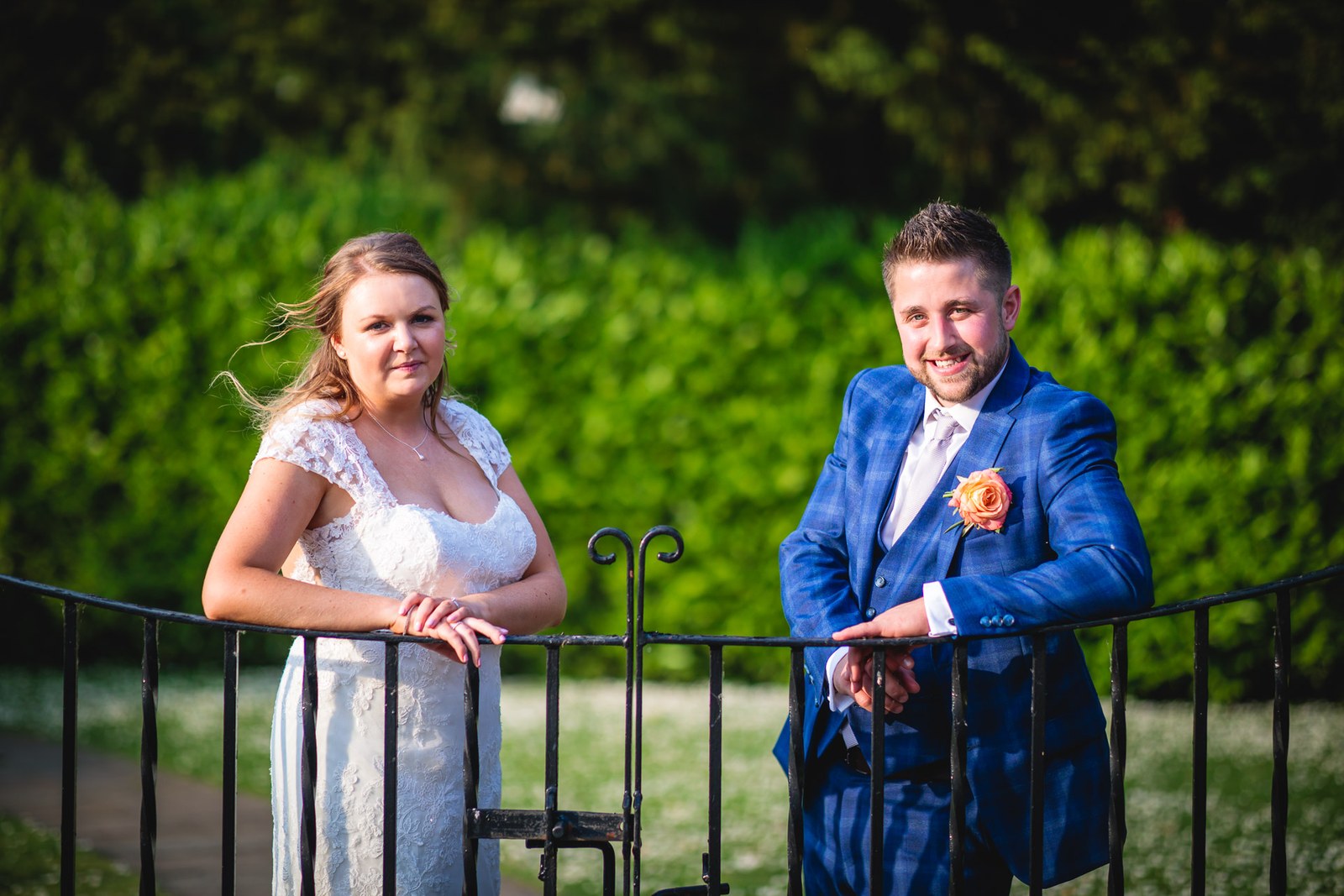 The couple resting on the black iron garden gate with a hedge behind