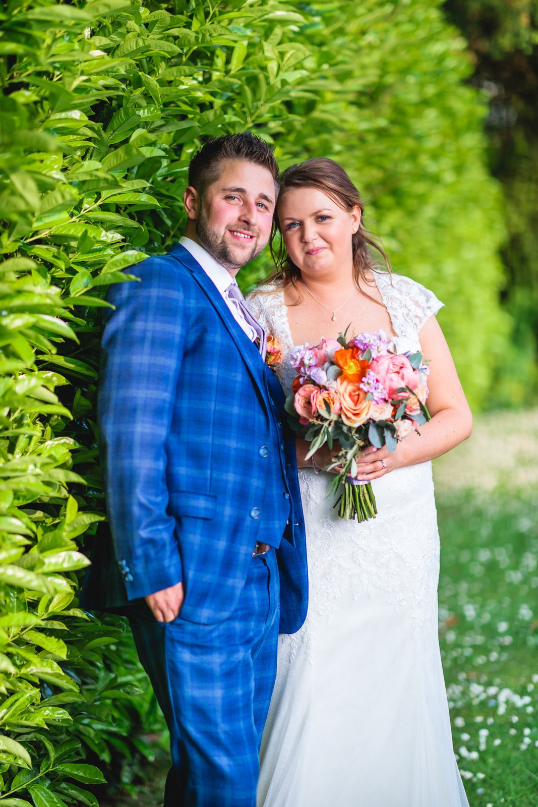 Bride and groom beside a tall green hedge, bride holding her coral bouquet