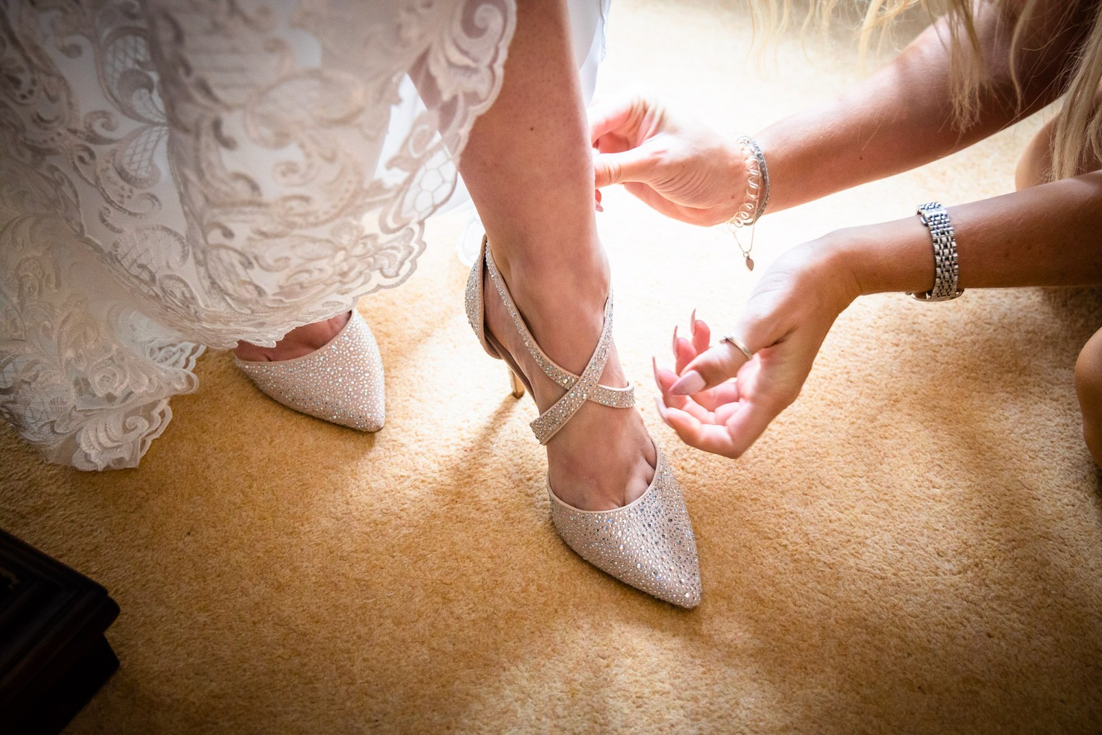 A bridesmaid fastening the strap of the bride's sparkly shoe below the lace hem